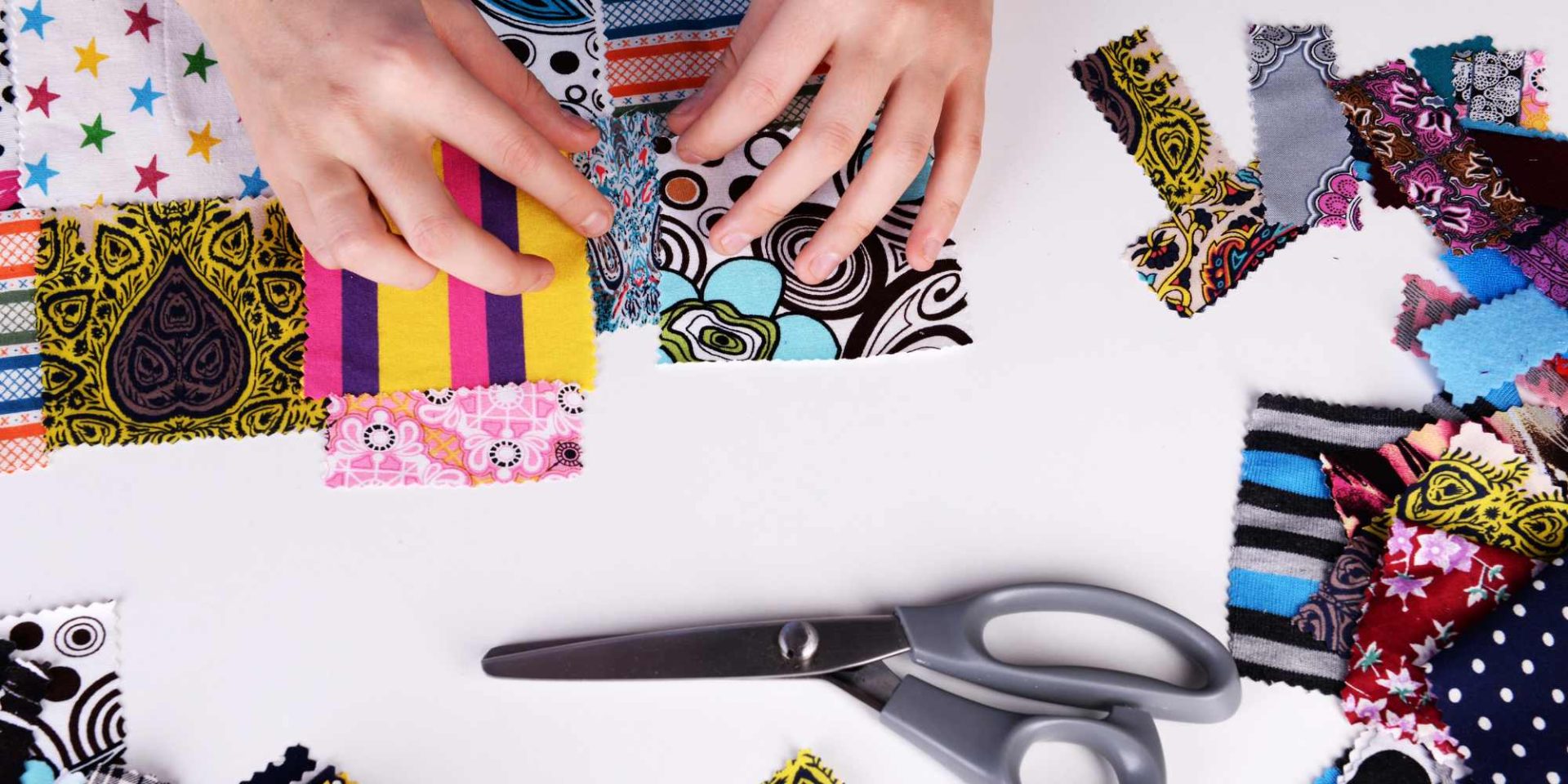 A close-up lifestyle image of hands sewing colourful patchwork fabric pieces together on a wooden table.