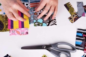 A close-up lifestyle image of hands sewing colourful patchwork fabric pieces together on a wooden table.